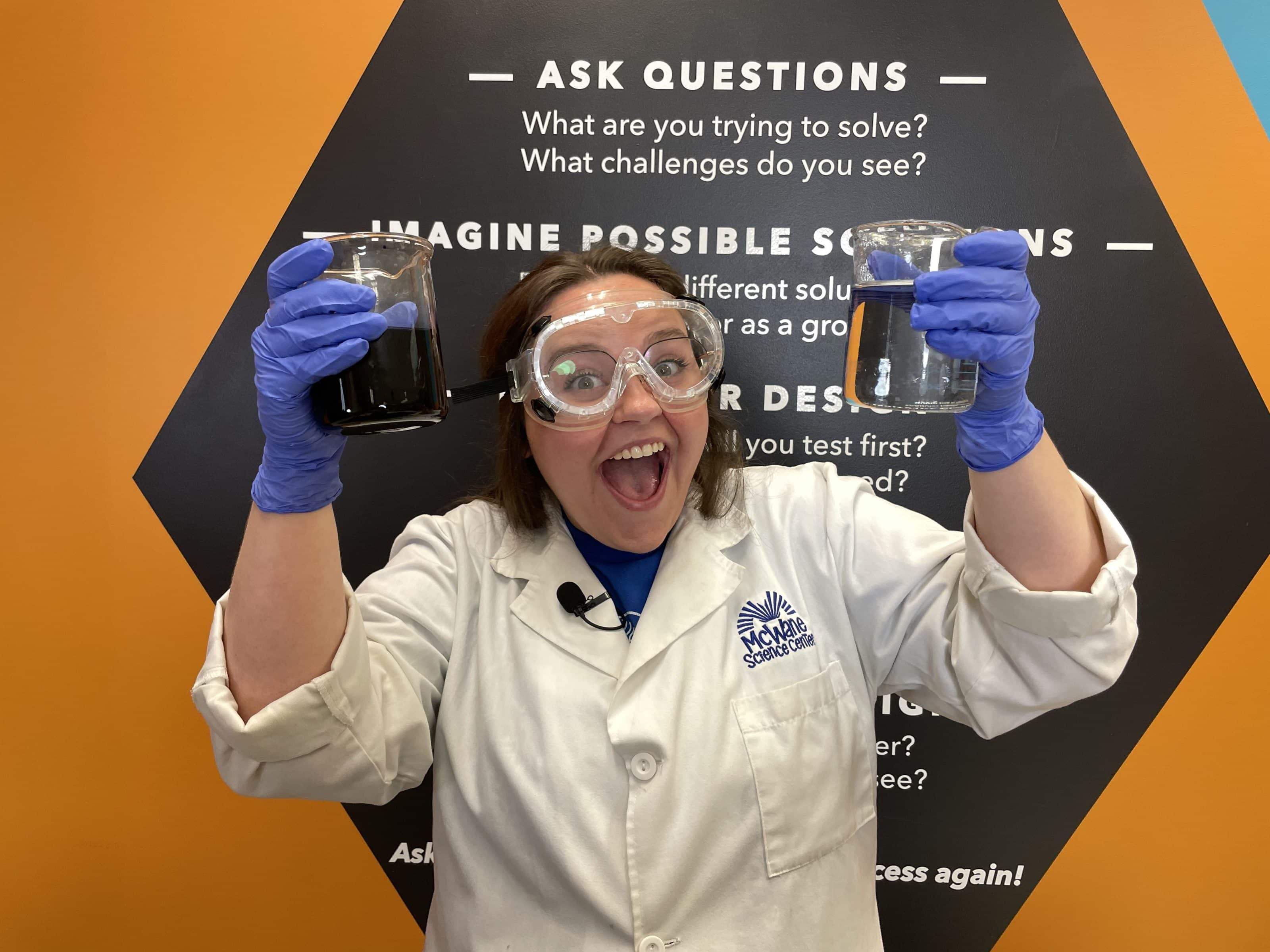 McWane Science Center educator in goggles holds beakers of dark and clear liquid, smiling during a hands-on STEM demo